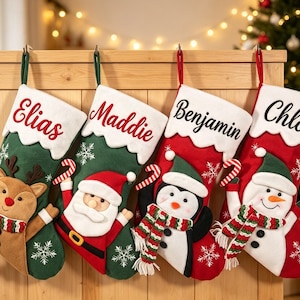 Op de afbeelding: Vier kerstsokken hangen op een rij. De eerste is groen met een rendier, de tweede groen met de Kerstman, de derde rood met een pinguïn en de vierde rood met een sneeuwpop. Elke sok heeft een witte boord met een naam erop geborduurd.