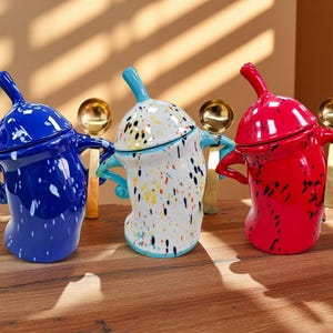 Puede incluir: Tres tarros de cerámica en azul, blanco y rojo, cada uno con un diseño de pintura moteada único. Los tarros tienen forma curva, asas pequeñas y una tapa con un asa larga y curva. Cucharas doradas se colocan detrás de cada tarro.