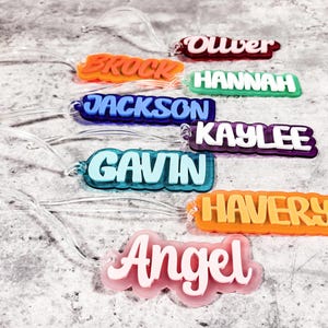 Może przedstawiać: Kolekcja kolorowych identyfikator&oacute;w z imionami z przezroczystymi plastikowymi pętelkami. Imiona to "Oliver", "Brock", "Hannah", "Jackson", "Kaylee", "Gavin", "Haverly" i "Angel". Każdy identyfikator ma inną kombinację kolor&oacute;w z białymi literami.