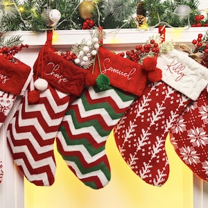 Op de afbeelding: Vijf kerstsokken in rood en wit, met geborduurde namen op de manchetten. Ontwerpen omvatten rendieren, sneeuwvlokken en zigzagpatronen. Elke sok heeft een pompon en hangt aan een witte schoorsteenmantel.