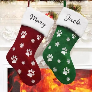 May include: Two Christmas stockings, one deep red and one forest green, each featuring a white faux fur cuff. The red stocking is embroidered with the name Mary, and the green stocking with Jack. Both have white paw prints and snowflake patterns.
