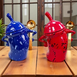 Puede incluir: Dos soperas de cerámica, una azul con motas blancas y otra roja con motas negras, cada una con tapa a juego y un cucharón dorado. Las soperas están sobre una mesa de madera.