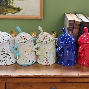 May include: Five ceramic jars with lids, each with a distinct design. The jars are white, blue, and red, with speckled paint patterns in various colours. They have handles and a spout-like top. The jars are displayed on a wooden surface.