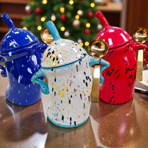 Puede incluir: Tres tarros de cerámica en azul, rojo y blanco con diseños de pintura salpicada colorida. Cada tarro tiene una tapa, un asa y una cuchara dorada. Los tarros están sobre una superficie marrón, con un árbol de Navidad al fondo.