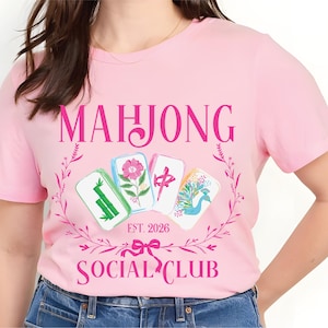 Puede incluir: Camiseta rosa claro con la inscripci&oacute;n "MAHJONG SOCIAL CLUB" en una fuente decorativa, y "EST. 2026" debajo. El dise&ntilde;o presenta cuatro fichas de Mahjong con motivos florales y de aves, rodeadas por una corona floral rosa.