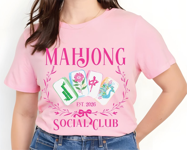 Puede incluir: Camiseta rosa claro con la inscripci&oacute;n "MAHJONG SOCIAL CLUB" en una fuente decorativa, y "EST. 2026" debajo. El dise&ntilde;o presenta cuatro fichas de Mahjong con motivos florales y de aves, rodeadas por una corona floral rosa.