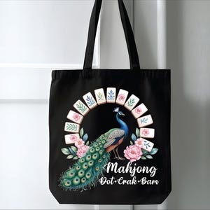 May include: Black tote bag featuring a colorful peacock design with a Mahjong theme. The peacock is surrounded by pink flowers and Mahjong tiles. The text "Mahjong Dot-Crak-Bam" is written in white script.
