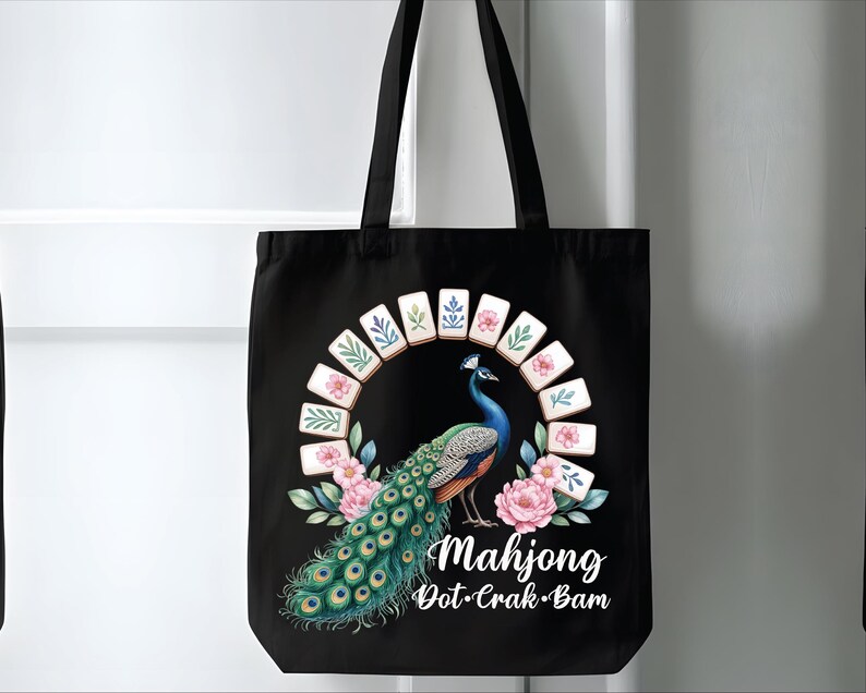 May include: Black tote bag featuring a colorful peacock design with a Mahjong theme. The peacock is surrounded by pink flowers and Mahjong tiles. The text "Mahjong Dot-Crak-Bam" is written in white script.