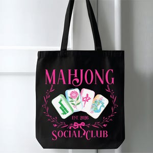 Puede incluir: Bolso tote negro con texto e ilustraciones rosas. Las palabras "MAHJONG SOCIAL CLUB" se muestran, junto con "EST. 2026". El dise&ntilde;o incluye cuatro fichas de Mahjong y una corona floral.