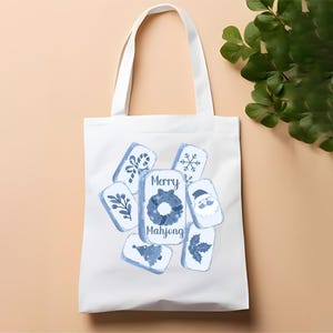 May include: White tote bag featuring blue and white illustrations of Mahjong tiles with holiday-themed designs. The text "Merry Mahjong" is displayed in the center. The bag has two white handles.