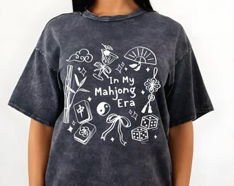 May include: Dark gray t-shirt with a distressed finish. The shirt features a white graphic design with the text "In My Mahjong Era" and illustrations of mahjong tiles, dice, and other related symbols.