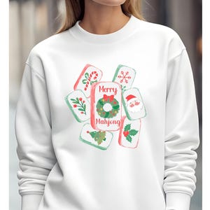 May include: White sweatshirt featuring a festive "Merry Mahjong" design. The design includes watercolor-style illustrations of Christmas-themed Mahjong tiles with candy canes, holly, a Christmas tree, and Santa Claus. The text "Merry Mahjong" is displayed prominently.
