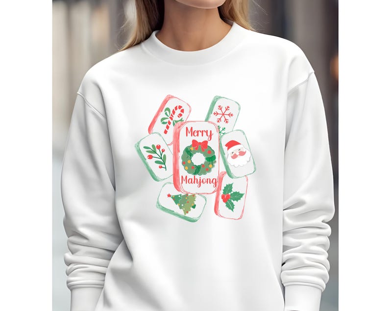 May include: White sweatshirt featuring a festive "Merry Mahjong" design. The design includes watercolor-style illustrations of Christmas-themed Mahjong tiles with candy canes, holly, a Christmas tree, and Santa Claus. The text "Merry Mahjong" is displayed prominently.