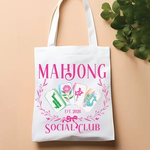 Puede incluir: Bolso tote blanco con texto rosa "MAHJONG SOCIAL CLUB EST. 2026". El bolso presenta un dise&ntilde;o floral e ilustraciones de fichas de Mahjong. El bolso tiene asas blancas y est&aacute; sobre un fondo melocot&oacute;n claro.