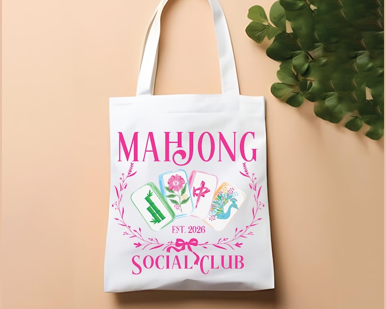 Puede incluir: Bolso tote blanco con texto rosa "MAHJONG SOCIAL CLUB EST. 2026". El bolso presenta un dise&ntilde;o floral e ilustraciones de fichas de Mahjong. El bolso tiene asas blancas y est&aacute; sobre un fondo melocot&oacute;n claro.