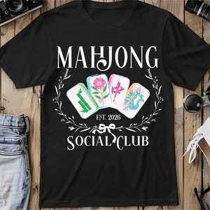 Puede incluir: Camiseta negra con la frase "MAHJONG SOCIAL CLUB" y el a&ntilde;o "EST. 2026". El dise&ntilde;o presenta cuatro fichas de Mahjong con ilustraciones florales y simb&oacute;licas, rodeadas por una corona floral blanca.