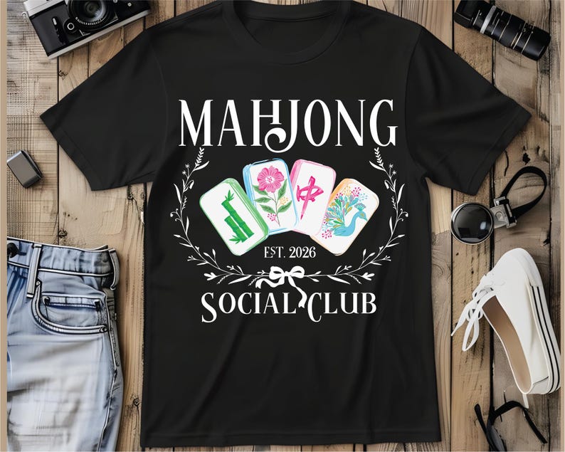 Puede incluir: Camiseta negra con la frase "MAHJONG SOCIAL CLUB" y el a&ntilde;o "EST. 2026". El dise&ntilde;o presenta cuatro fichas de Mahjong con ilustraciones florales y simb&oacute;licas, rodeadas por una corona floral blanca.