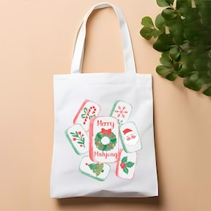 May include: White tote bag featuring a festive design. The bag displays colorful illustrations of Christmas-themed Mahjong tiles. The central tile reads "Merry Mahjong" with a wreath. Other tiles show a candy cane, snowflake, Santa, and more.