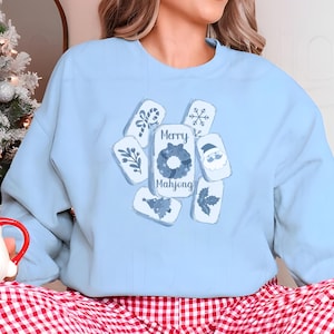 May include: A light blue sweatshirt featuring a festive design. The design includes several rectangular shapes with holiday-themed illustrations and the words "Merry Mahjong". A Christmas tree and a Santa mug are visible in the background.