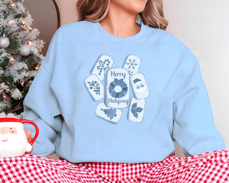 May include: A light blue sweatshirt featuring a festive design. The design includes several rectangular shapes with holiday-themed illustrations and the words "Merry Mahjong". A Christmas tree and a Santa mug are visible in the background.