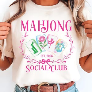 Puede incluir: Camiseta color crema con un dise&ntilde;o rosa. El dise&ntilde;o incluye la palabra "MAHJONG", una corona floral y cuatro fichas de Mahjong ilustradas. Debajo, el texto "SOCIAL CLUB" y "EST. 2026" se muestran en rosa. Es una prenda casual.