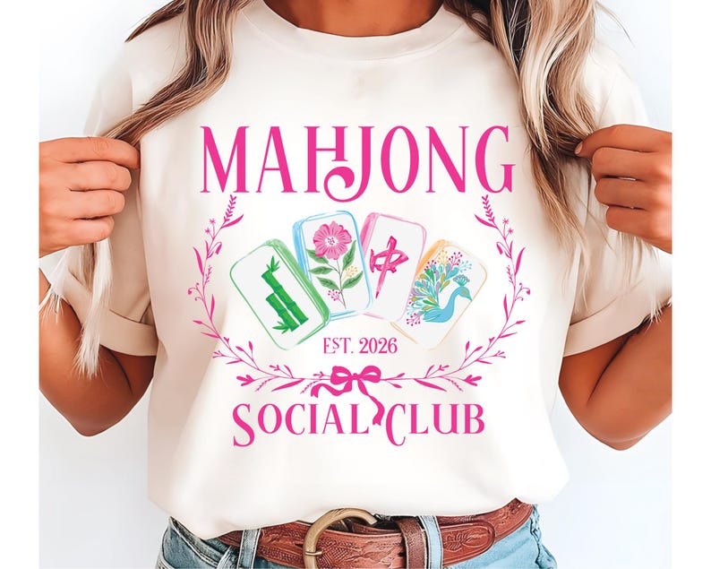 Puede incluir: Camiseta color crema con un dise&ntilde;o rosa. El dise&ntilde;o incluye la palabra "MAHJONG", una corona floral y cuatro fichas de Mahjong ilustradas. Debajo, el texto "SOCIAL CLUB" y "EST. 2026" se muestran en rosa. Es una prenda casual.