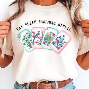 May include: A cream-colored t-shirt featuring a graphic of four mahjong tiles with floral, bird, and vase illustrations. Above the tiles, the text reads EAT. SLEEP. MAHJONG. REPEAT. in a black serif font. The shirt has short sleeves and a crew neckline.