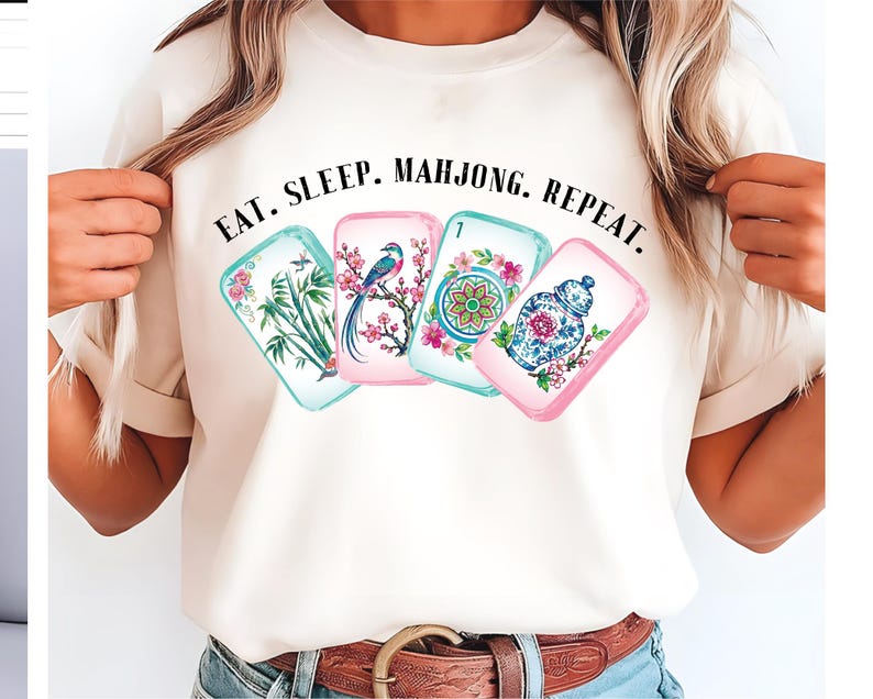 May include: A cream-colored t-shirt featuring a graphic of four mahjong tiles with floral, bird, and vase illustrations. Above the tiles, the text reads EAT. SLEEP. MAHJONG. REPEAT. in a black serif font. The shirt has short sleeves and a crew neckline.