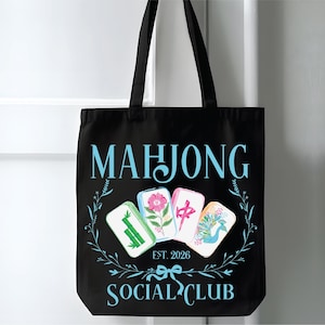 Puede incluir: Bolso tote negro con las palabras "MAHJONG SOCIAL CLUB" en azul claro. El bolso presenta cuatro fichas de Mahjong con dise&ntilde;os florales y s&iacute;mbolos chinos. Tambi&eacute;n se incluye el texto "EST. 2026".