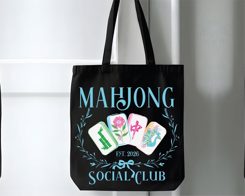 Puede incluir: Bolso tote negro con las palabras "MAHJONG SOCIAL CLUB" en azul claro. El bolso presenta cuatro fichas de Mahjong con dise&ntilde;os florales y s&iacute;mbolos chinos. Tambi&eacute;n se incluye el texto "EST. 2026".