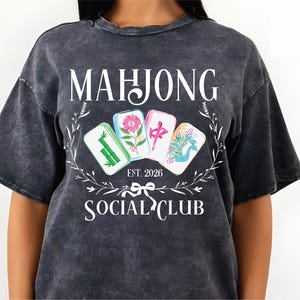 Puede incluir: Camiseta gris oscuro con las palabras "MAHJONG SOCIAL CLUB" y "EST. 2026" en blanco. La camiseta presenta un dise&ntilde;o floral y cuatro fichas de Mahjong coloridas. La camiseta est&aacute; hecha de un material suave y c&oacute;modo.