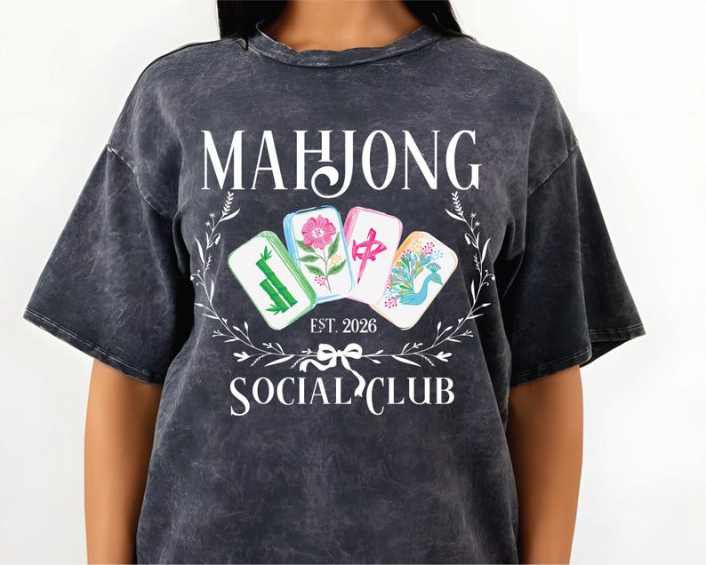 Puede incluir: Camiseta gris oscuro con las palabras "MAHJONG SOCIAL CLUB" y "EST. 2026" en blanco. La camiseta presenta un dise&ntilde;o floral y cuatro fichas de Mahjong coloridas. La camiseta est&aacute; hecha de un material suave y c&oacute;modo.