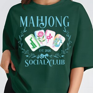 Puede incluir: Sudadera verde oscuro con las palabras "MAHJONG SOCIAL CLUB" en azul claro. El dise&ntilde;o incluye cuatro fichas de Mahjong con ilustraciones florales y de aves, y el texto "EST. 2026".