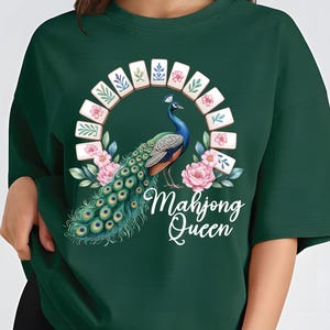May include: Dark green sweatshirt featuring a peacock design with a floral and tile arch. The peacock has blue, green, and orange feathers. The text "Mahjong Queen" is written in white script below the peacock.