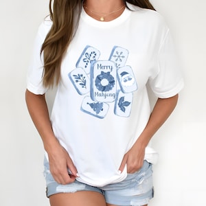 May include: White t-shirt featuring a blue watercolor design of Mahjong tiles with holiday-themed illustrations. The central tile reads "Merry Mahjong". The design includes a snowflake, Santa, and other festive elements.