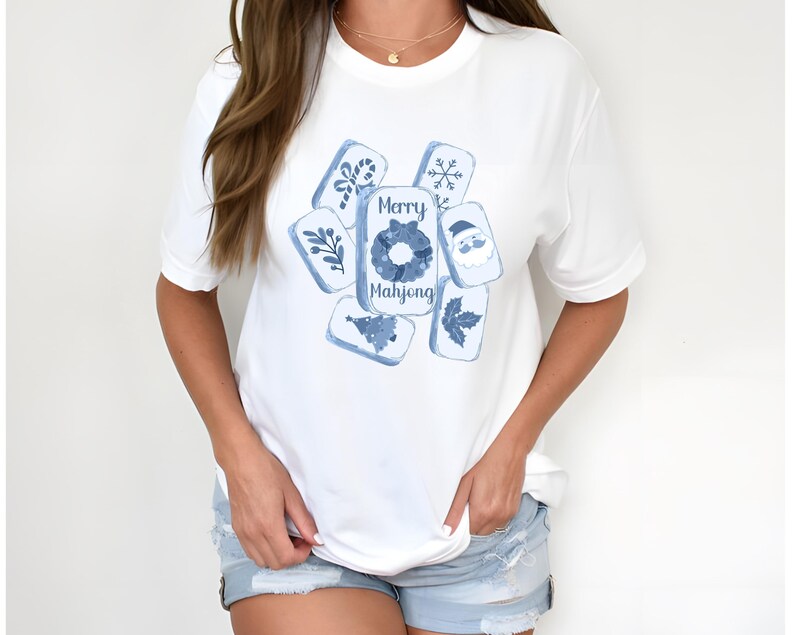 May include: White t-shirt featuring a blue watercolor design of Mahjong tiles with holiday-themed illustrations. The central tile reads "Merry Mahjong". The design includes a snowflake, Santa, and other festive elements.
