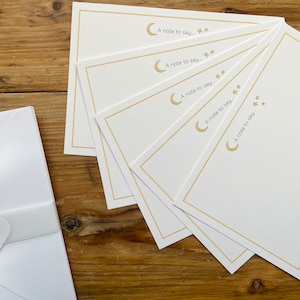 May include: A set of five white notecards with gold borders and a crescent moon and stars design. The notecards have the text "A note to say..." printed on them.