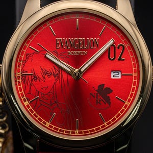 Pu&ograve; includere: Orologio da polso color oro con quadrante rosso con un'illustrazione di un personaggio anime e la scritta "EVANGELION BOXFUN". L'orologio ha lancette dorate e un datario. Il cinturino &egrave; nero.