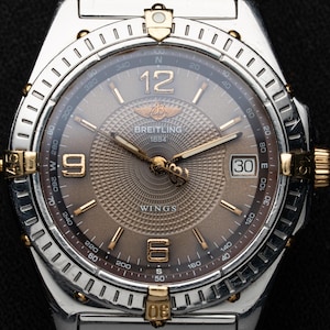 May include: A Breitling Wings wristwatch featuring a brown dial with a textured guilloché center, gold-tone hands, and gold-tone Arabic numerals at 6, 9, and 12 o'clock. The watch includes a date window at 3 o'clock and a rotating silver bezel.