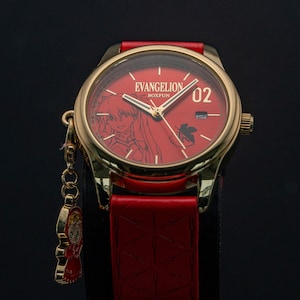 Pu&ograve; includere: Un orologio dorato con quadrante e cinturino rossi. Il quadrante mostra il testo "EVANGELION BOXFUN" e un'illustrazione di un personaggio. Un piccolo ciondolo &egrave; appeso all'orologio. Il cinturino &egrave; in pelle rossa.