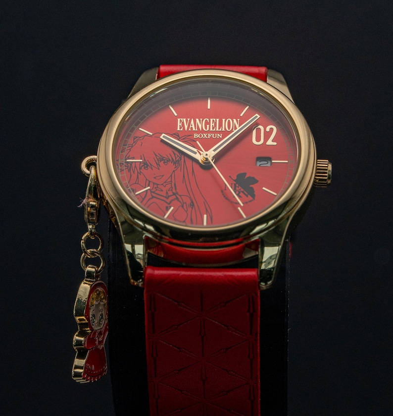 Pu&ograve; includere: Un orologio dorato con quadrante e cinturino rossi. Il quadrante mostra il testo "EVANGELION BOXFUN" e un'illustrazione di un personaggio. Un piccolo ciondolo &egrave; appeso all'orologio. Il cinturino &egrave; in pelle rossa.