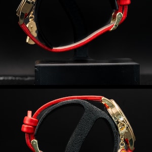 Pu&ograve; includere: Orologio da polso dorato con cinturino in pelle rosso. L'orologio ha un quadrante dorato e un piccolo ciondolo di una ragazza con un vestito rosso. L'orologio &egrave; esposto su un supporto nero su sfondo scuro.