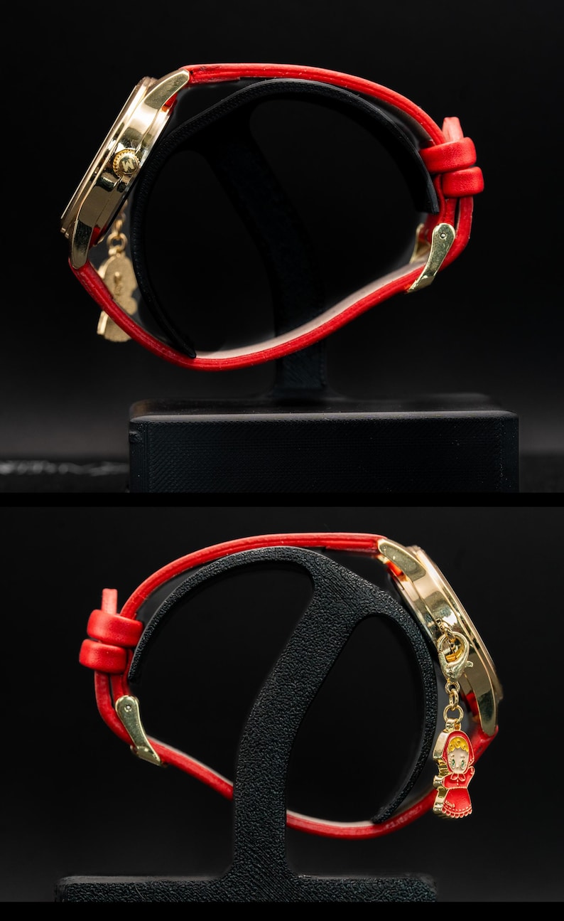 Pu&ograve; includere: Orologio da polso dorato con cinturino in pelle rosso. L'orologio ha un quadrante dorato e un piccolo ciondolo di una ragazza con un vestito rosso. L'orologio &egrave; esposto su un supporto nero su sfondo scuro.