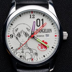 Pu&ograve; includere: Orologio da polso color argento con quadrante bianco con illustrazione di un personaggio anime e la scritta "EVANGELION". L'orologio ha lancette nere con dettagli rossi e verdi, cinturino in pelle nera e datario.