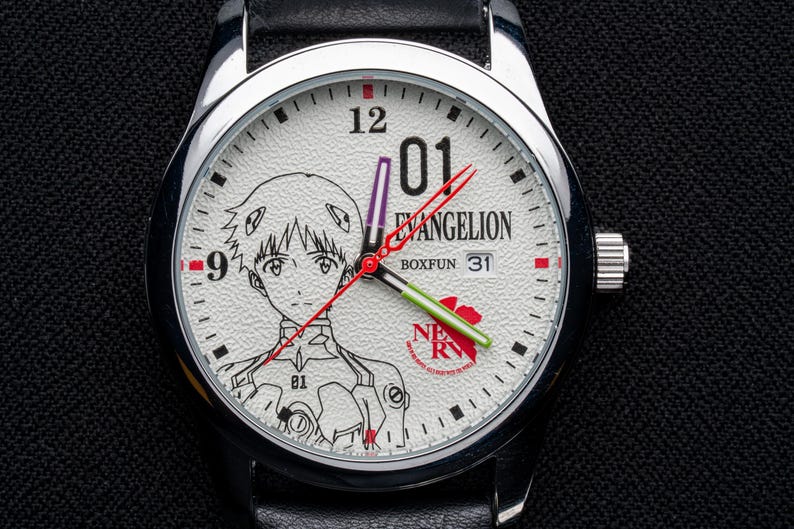Pu&ograve; includere: Orologio da polso color argento con quadrante bianco con illustrazione di un personaggio anime e la scritta "EVANGELION". L'orologio ha lancette nere con dettagli rossi e verdi, cinturino in pelle nera e datario.
