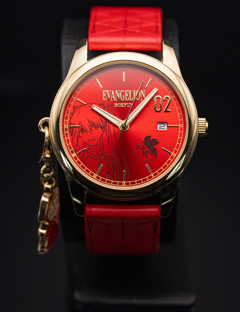 May include: A gold-toned wristwatch with a red face and strap. The watch face features the text "EVANGELION" and a character illustration. The strap is red leather. A small charm hangs from the watch. The watch is displayed on a black surface.