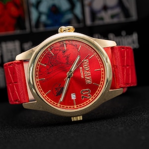 Pu&ograve; includere: Un orologio dorato con quadrante rosso e cinturino in pelle rosso. Il quadrante dell'orologio presenta un design grafico e il testo "EVANGELION BOSTON 02". L'orologio ha lancette dorate e una corona dorata.