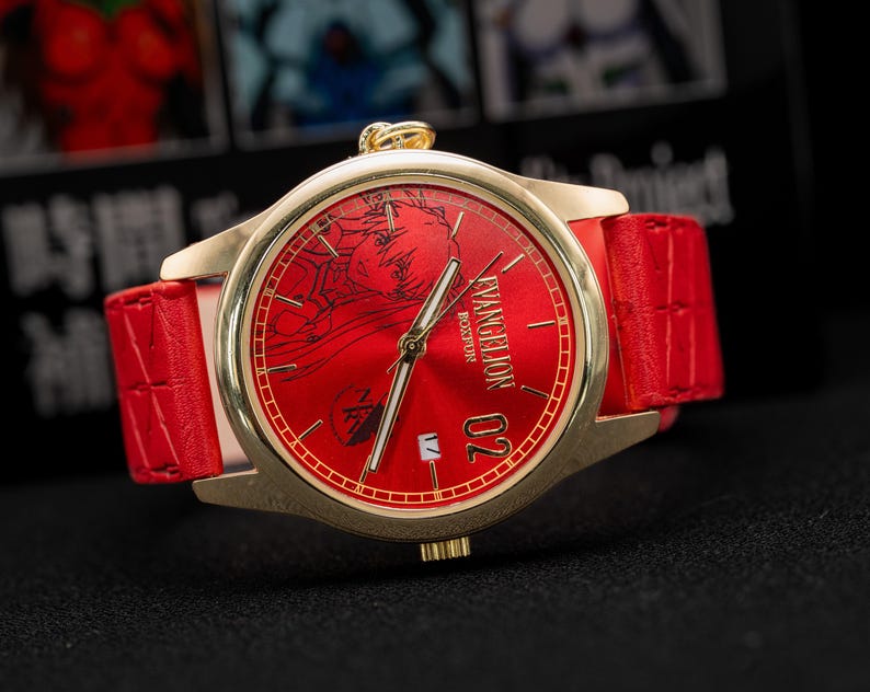 Pu&ograve; includere: Un orologio dorato con quadrante rosso e cinturino in pelle rosso. Il quadrante dell'orologio presenta un design grafico e il testo "EVANGELION BOSTON 02". L'orologio ha lancette dorate e una corona dorata.