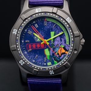 Pu&ograve; includere: Un orologio da polso grigio scuro con cinturino in nylon viola. Il quadrante blu presenta una grafica di un personaggio viola e verde. Le lancette dell'orologio sono rosse e verdi. Il testo "非常事態" &egrave; visibile.