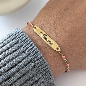 Poignet portant un bracelet personnalisé. C'est un bracelet type gourmette en acier inoxydable avec une chaine de couleur or et des perles roses. Un rectangle est gravé par un prénom ''Marine" d'une écriture cursive.